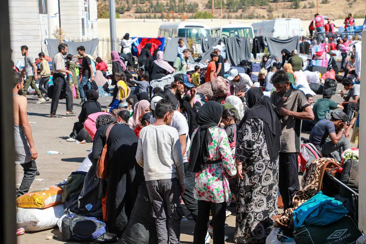 Des familles se tiennent debout et s'assoient sur une route pavée de terre avec de nombreux objets personnels. Des véhicules et des travailleurs humanitaires apparaissent à l'arrière-plan et des couvertures sont suspendues à des cordes pour sécher.