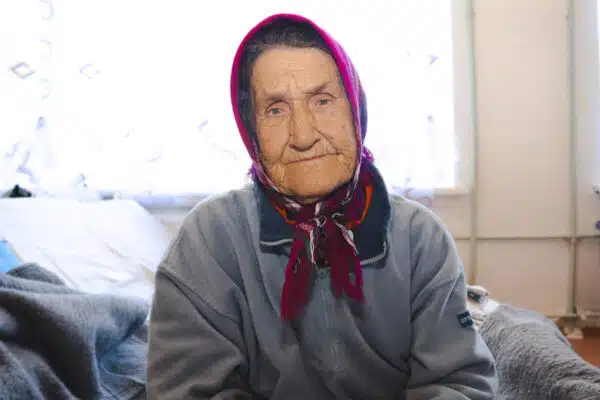 Woman wearing a pink and blue headcovering that ties at the bottom, stares at the camera while sitting on the edge of a bed.