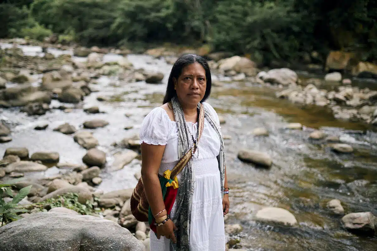 A person looks left at the camera which is slightly to their right, as their body faces further right. They wear a white dress and colourful bag while stood in front of a low river.