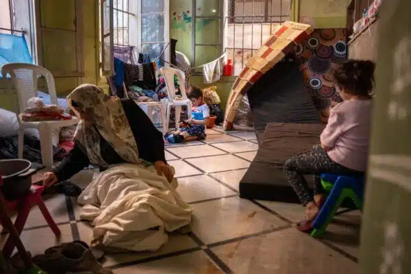 Deux enfants sont assis dans une pièce et s'occupent avec des jouets. Une femme âgée est agenouillée à côté d'une cuisinière portable et porte un couvre-chef.