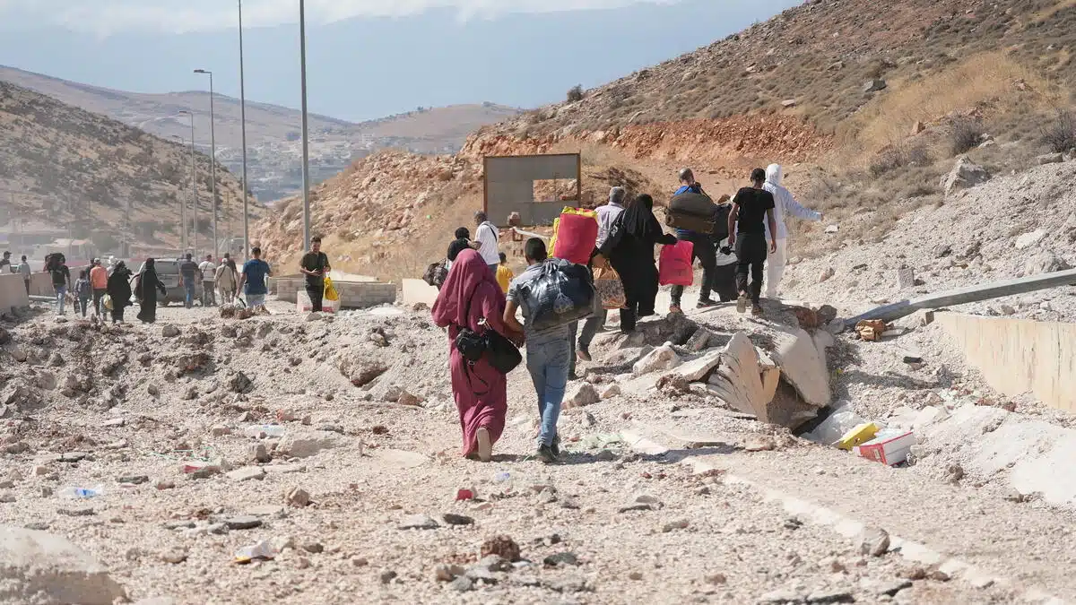 At least 26 people carry their belongings as they walk down a road filled with rubble. The road is surrounded by mounds of dirt and sand, with grass being sparse.