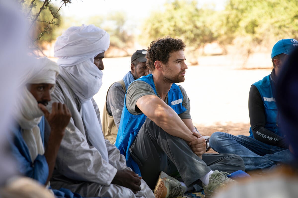 Person sits with their arm over their propped up leg, wearing a blue UNHCR vest. Another person in a UNHCR vest sits on his right. Two people sit on his left in a white headcovering.