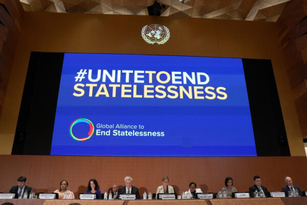 Un écran situé au-dessus de plusieurs personnes assises à un bureau face à la caméra affiche « #UNITEDTOENDSTATELESSNESS » ainsi que le logo de l'Alliance mondiale pour mettre fin à l'apatridie.