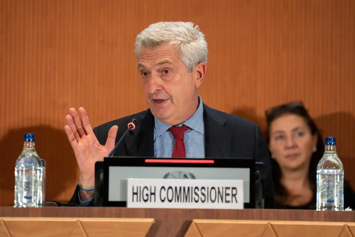 Man waves hand while sat behind a panel displaying "HIGH COMMISSIONER". Water bottles sit on the table and a woman sits in the background.