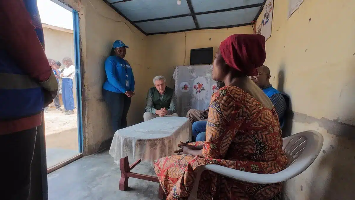 Two people in blue UNHCR vests sit in a room on each side, furthest from the camera. A man sits with his hands resting on his knees, bending forward so as to listen better. A woman in a red head covering and orange dress speaks to him while sitting in a law chair. Another person wearing a UNHCR vest stands to the left of the camera, blurred.