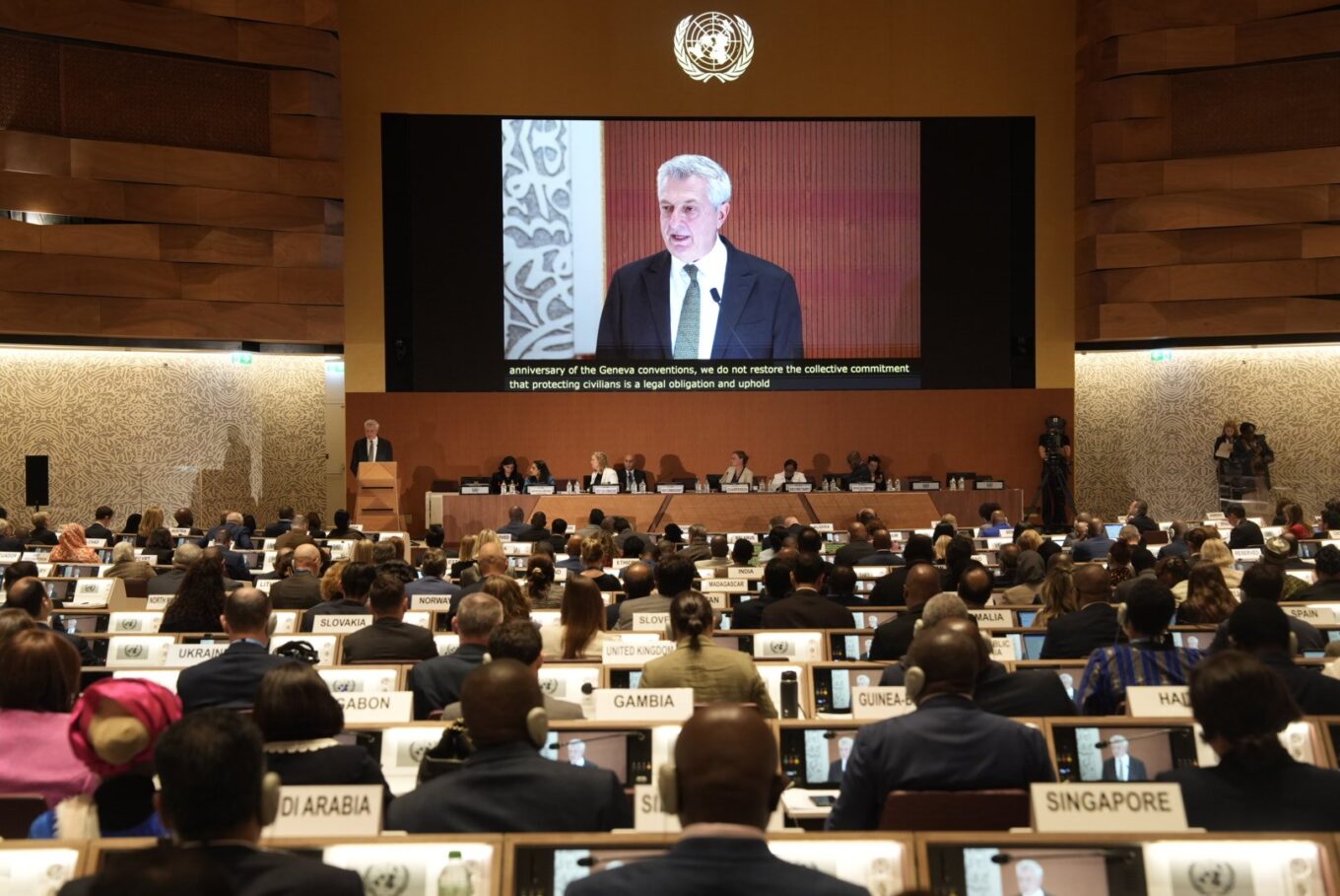 A room filled with formally dressed country representatives face towards the front of the room where a screen displays the speaker, Filippo Grandi who is speaking at the front of the room. People sit behind a table next to the speaker at the front of the room.