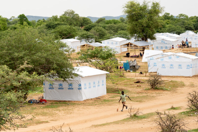 Shelters line a dirt road which display the blue UNHCR logo on a white background. A boy sprints down the road while carrying a stick-like toy.