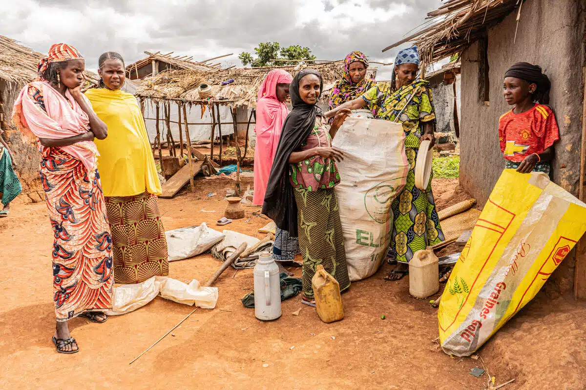 Two women on the left are slightly separate from the group on the right. They are wearing long and colourful cultural dresses.Four women on the right in colourful cultural clothing pack belongings into a bag. A young boy stands to their right and gazes across the screen while holding a yellow bag. They stand on a dirt patch and shelters can be seen around.