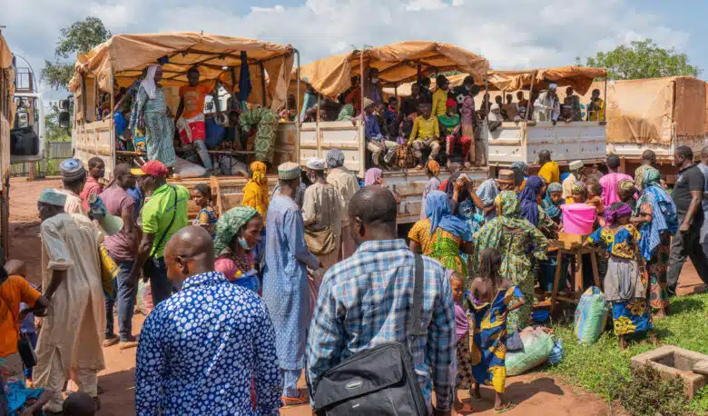 Four large caravan trucks are parked with their rear facing the camera. The three in the center are filled with several people wearing colourful clothing. More people stand with their families at the bottom of the rear of the trucks. 