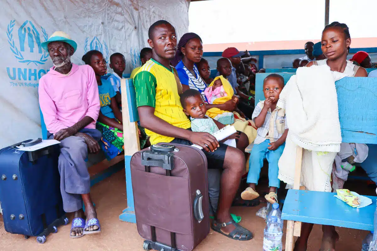 Several people sit lined up in a a canopy-like shelter. They hold documents and carry suitcases with them as some glance toward the direction of the camera.