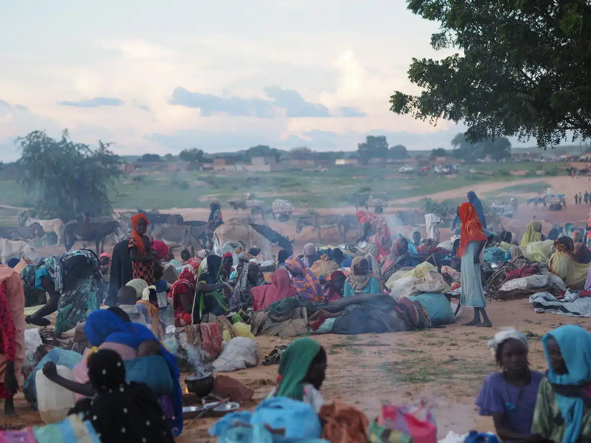 Several groups gather in circle shapes on a dusty part of land, the land nearest to them is green and their horses graze there. Smoke fills the air from fires that each are using to cook.