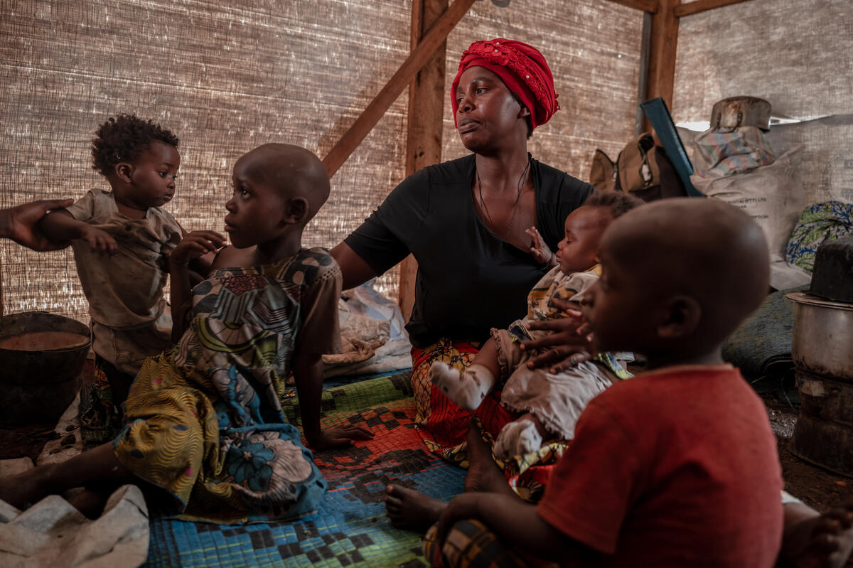 Une femme est assise sur un tapis à motifs, tenant un enfant dans ses bras tandis qu'une personne hors champ lui en passe un autre. Deux enfants plus âgés sont assis avec elle.