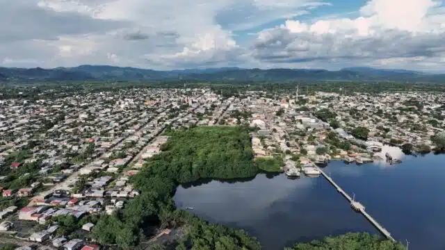 A city with a forest and body of water in the center and bottom right of the image is pictured from an aerial view. a bridge runs across the water in the bottom right. The environment is filled by clouds in the sky and mountains in the background.