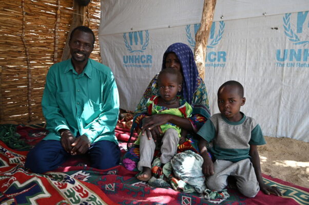 Une famille est assise à genoux à l'intérieur d'une tente dont les murs affichent le logo du HCR. Le sol en sable est recouvert d'une couverture colorée et à motifs sur laquelle ils s'assoient. Un homme sourit à l'extrême gauche, une femme regarde vers le bas tout en tenant un enfant, l'enfant tenu regarde la caméra et un enfant à l'extrême gauche regarde également la caméra.