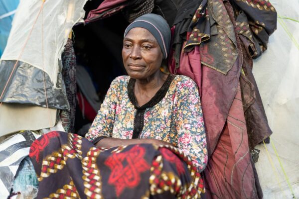 A woman stands blankly in front of her.She is wearing a floral blouse and patterned skirt. She sits with her knees folded into her chest, hands in her lap in front of an entrance to a tent shelter.