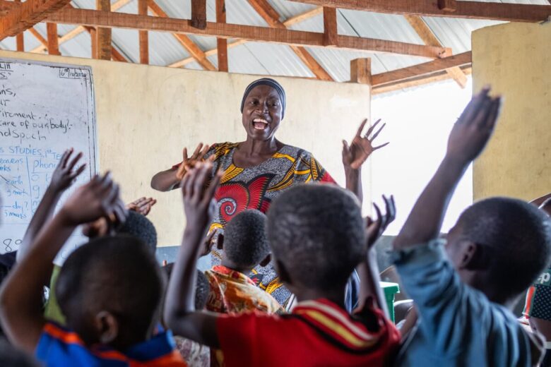 Woman expressively sways her hands in the air while smiling towards a group of children. The children face her, away from the camera.