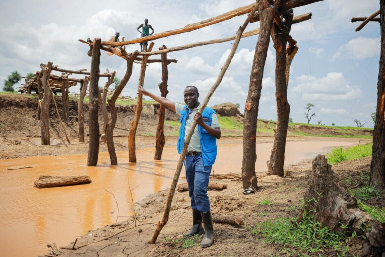 Un homme portant un gilet bleu ouvert tend le bras vers un objet situé à l'extérieur de l'image, sur le côté gauche. La personne semble plus basse que le début d'un humble pont en bois au-dessus d'elle. Il se tient à côté d'une eau boueuse et tient un grand bâton dans ses mains.