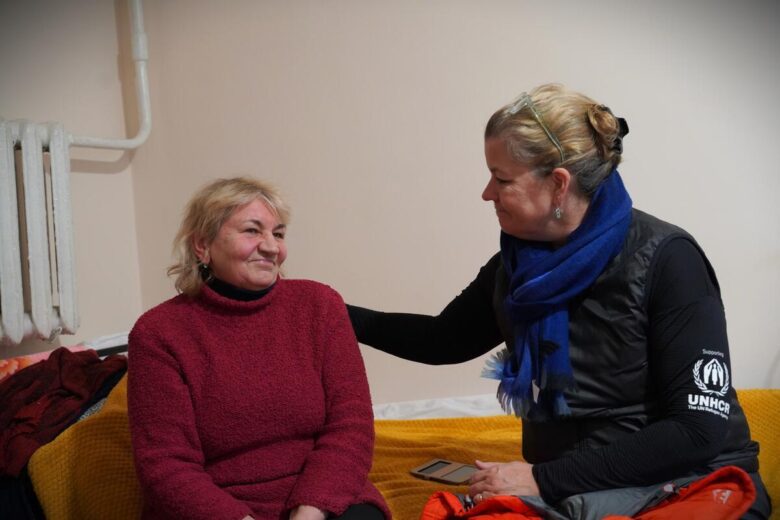 Une femme réconforte une autre femme alors qu'elles sont assises ensemble sur un lit. La femme qui réconforte l'autre porte une chemise à manches longues du HCR. La femme réconfortée porte un pull rouge.