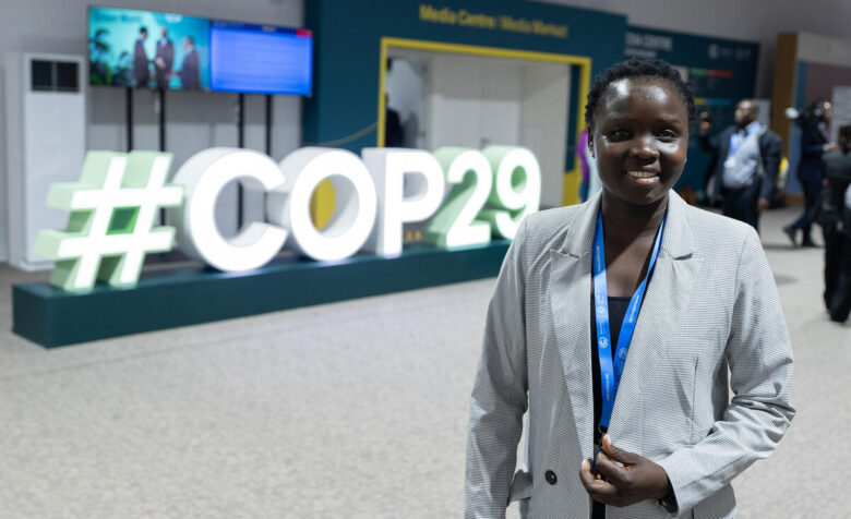 Une personne se tient devant un grand écran affichant « #COP29 », faiblement éclairé par des teintes vertes et jaunes. La personne sourit et tire sur son blazer pour le fermer.