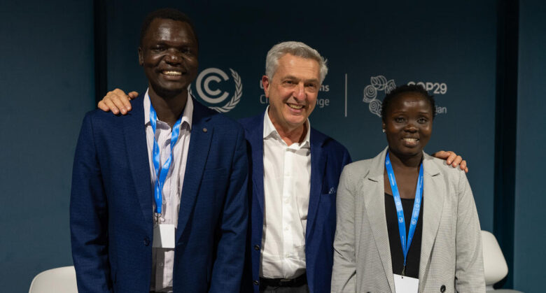 Trois personnes se tiennent de gauche à droite, de la plus grande à la moins grande. La personne du milieu rejoint les deux personnes sur les côtés en affichant un large sourire et en posant une main sur chacune de leurs épaules.