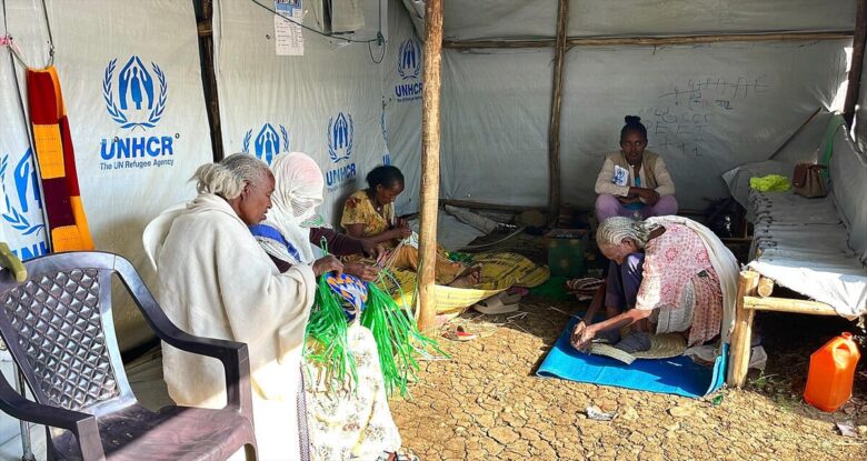 Five people sit in a shelter. Four of them are weaving material and the fifth is watching them.