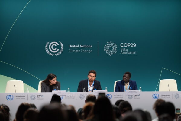 Three people sit at a panel in front of an emerald green backdroup which displays the logo for United Nations Climate Change and COP29. The two people on the outside look inward while the person in the middle gazes into the crowd beneath them.