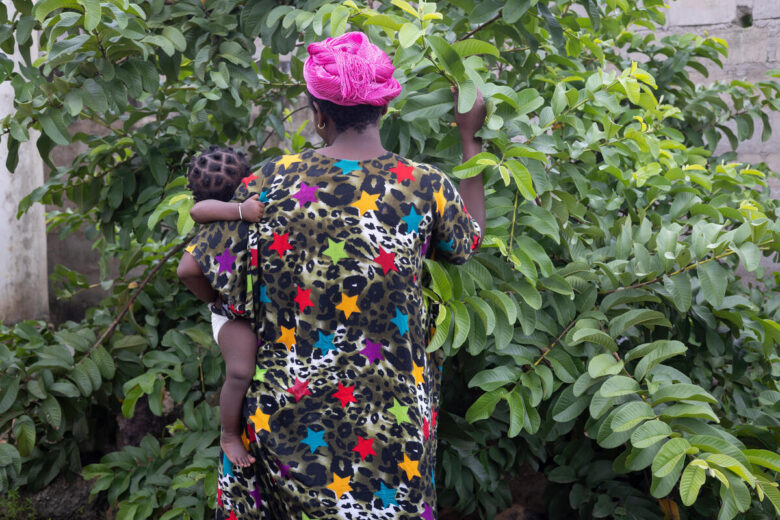 A woman, with her back to us, holds a baby in her arms while they observe and touch the leaves of a tree. She wears a black dress with colorful stars and a pink scarf on her head.