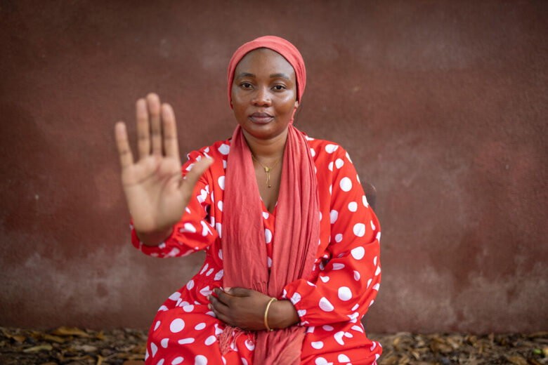 A woman wearing a red dress with white polka dots and a red scarf on her head is seated, holding one hand up in a "stop" gesture, while her other hand rests on her lap.