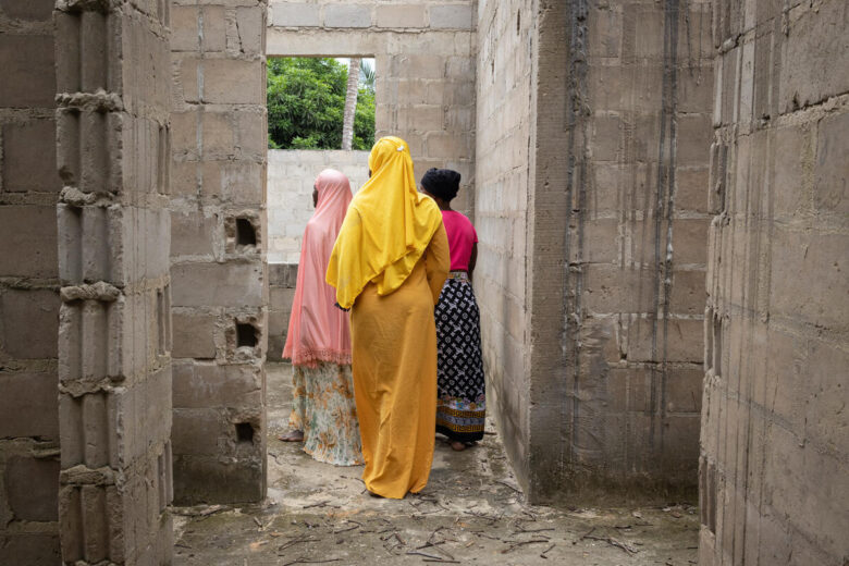 Three women walk through an abandoned building. They wear colorful yellow and pink tunics 