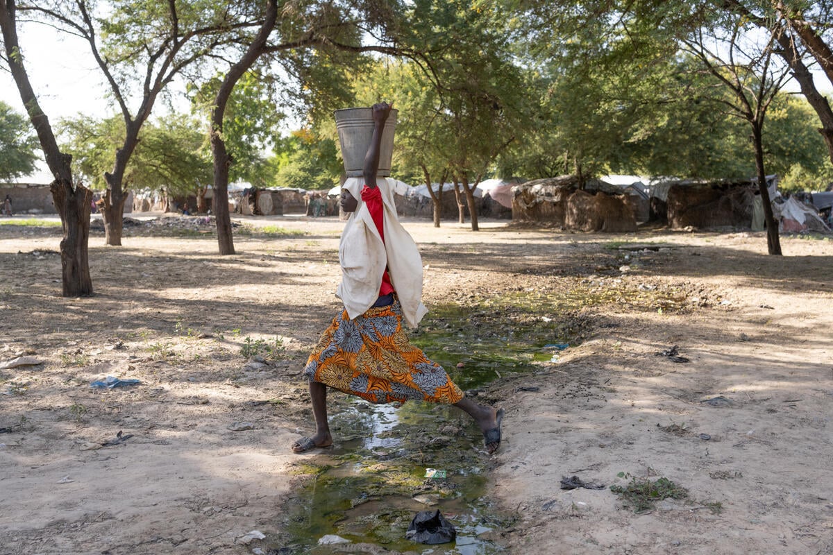 Woman carrying metal bucket on head, wearing white head scarf. She is wearing a red shirt and orange patterned skirt. The ground is light brown with trees in the back.