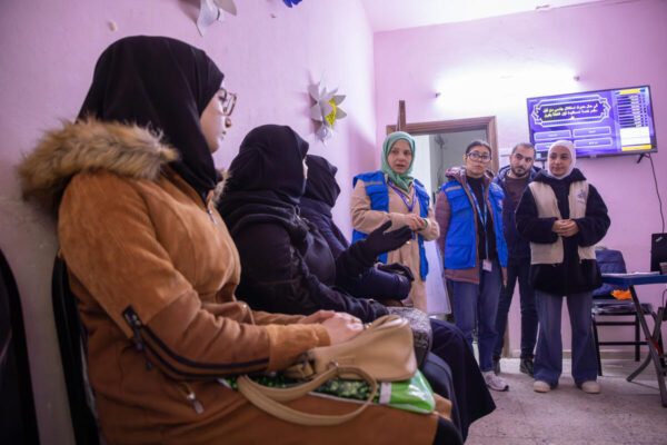 A group of women wearing black hijabs and winter coats sit in a waiting room in a community centre while three other women and a man standing by the door and wearing UNHCR blue jackets talk to them.