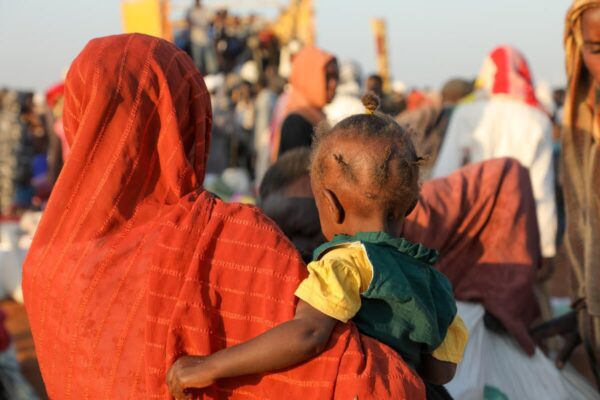 A photo of a woman with her back to the camera. She wears an orange headscarf over her head and shoulders while holding in her arms a small girl in a bright yellow dress and her hair braded into short braids.