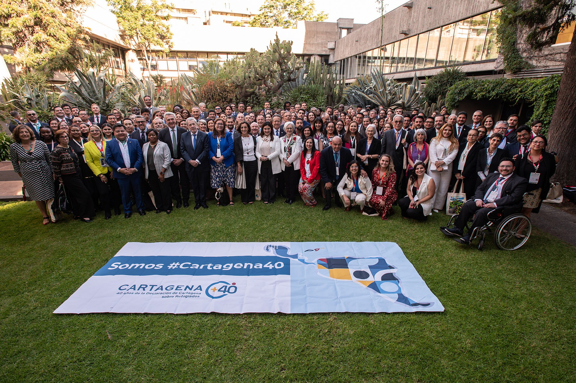 A large group of people pose for a photo on a green lawn with a large banner of the Cartagena+40 event visible in the foreground