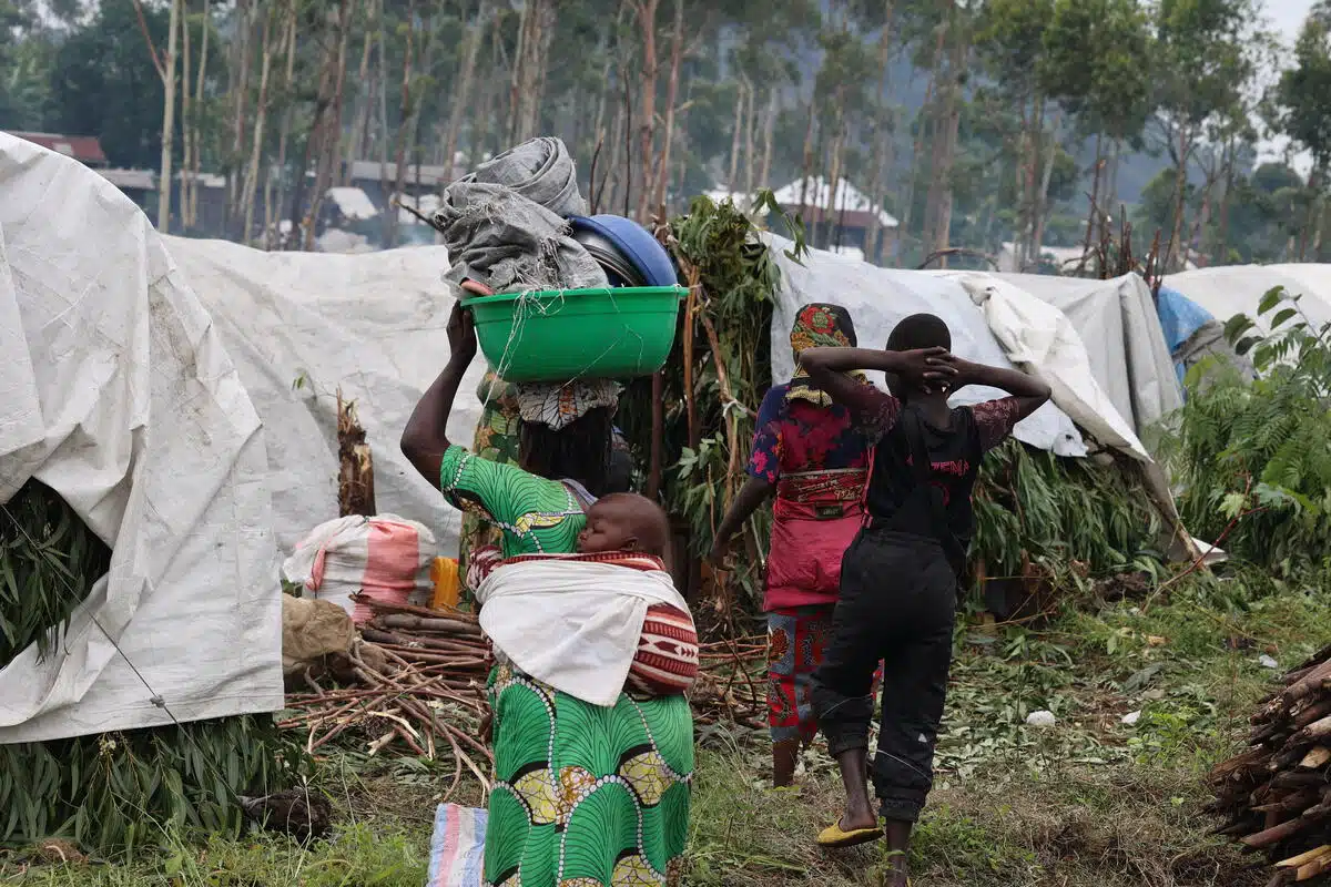 A woman dressed in a bright green dress with a small baby tied to her back carries on her head a large, green basin filled with other items. In the background there are makeshift shelters made of branches and covered with white tarps.