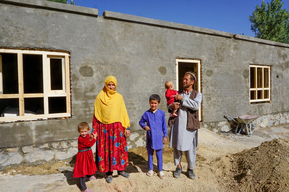 A family with their three children is standing outside the construction of their new house. The mother is wearing a red dress with blue flowers and a yellow headscarf covering her head. Her young daughter, holding her hand, is dressed in a red dress. The son is wearing a purple shirt and pants. The father is wearing a gray shirt and pants with a brown vest, holding their baby in his arms. The setting is outdoors near a house under construction, with a simple and natural background.