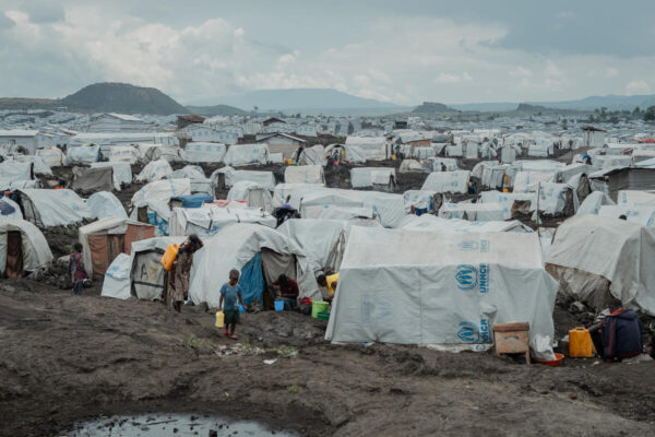 A young child walks across muddy ground in front of white hundreds of UNHCR tents pitched on a plain with mountains visible in the far distance.