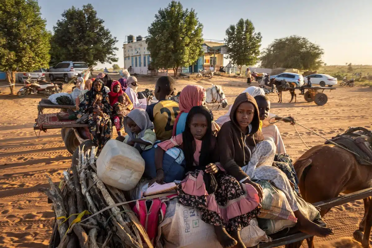 A Sudanese family of seven packed into the back of a horse-drawn carriage with their belongings ride on a dusty road with other families seen in the background.