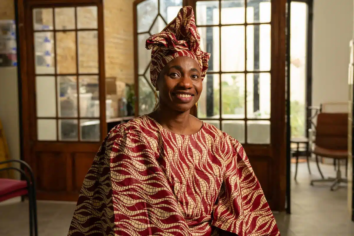 A woman is sitting in an office, smiling. She is wearing a brown and white dress with a matching headscarf.