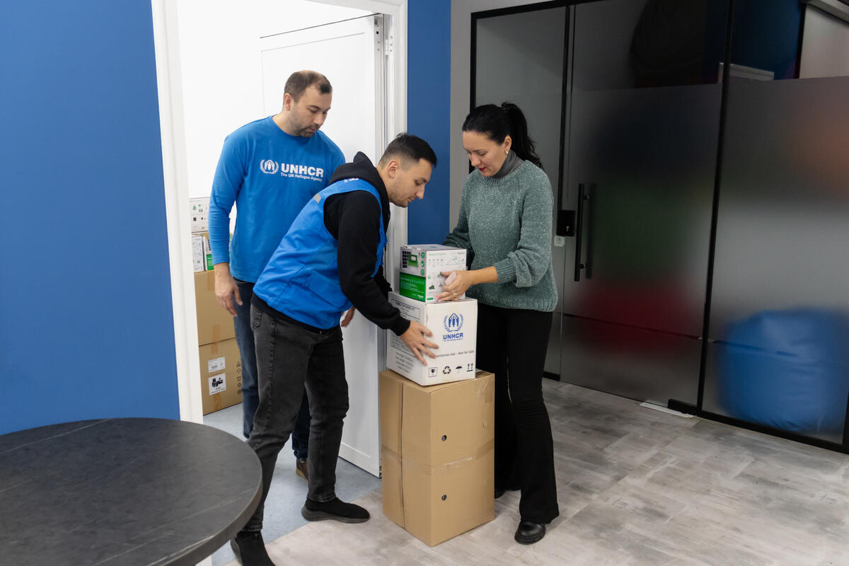 Three people from UNHCR are organizing boxes inside an office. There's a large container behind them