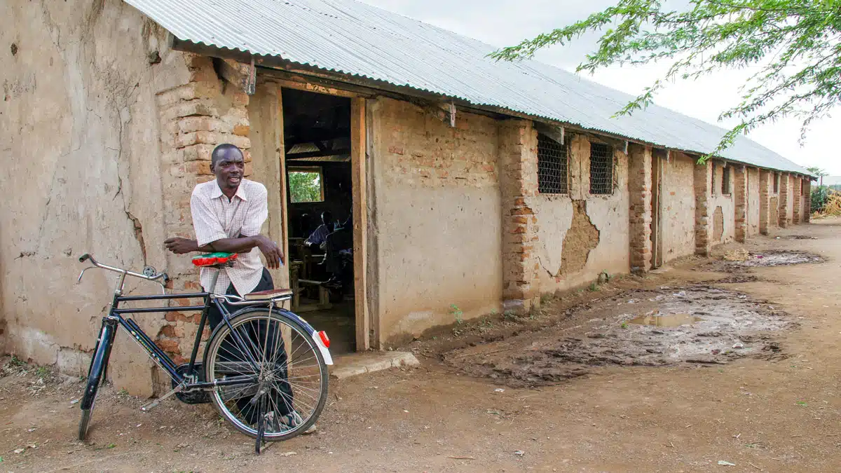 A man is leaning on his bike, wearing a white shirt and black pants. Behind him, there's a brick structure, and the ground is dirt.