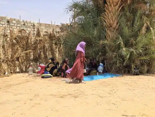 A young girl wearing a pink hijab and a long red dress walks by a group of women and children sitting on the sand in the shadow of palm trees.