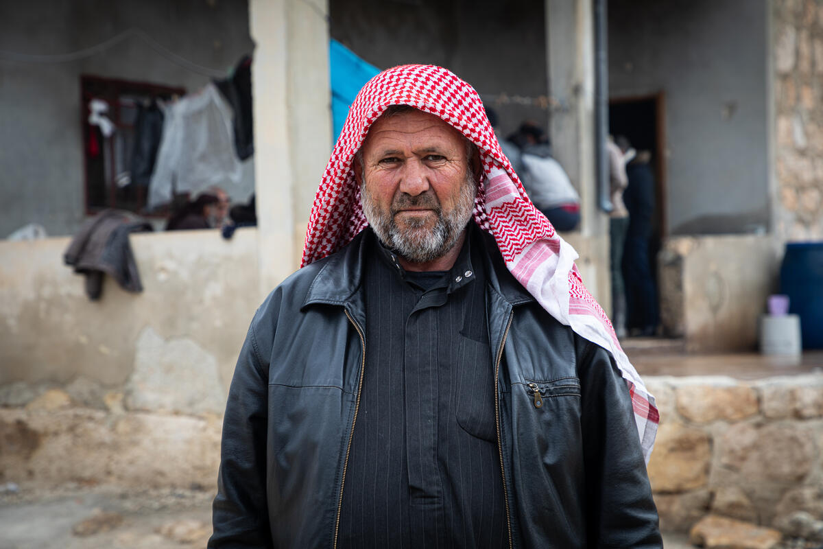 A man with a grey beard, dressed in black and wearing a red scarf on his head, stands outside a house under construction, looking directly at the camera.