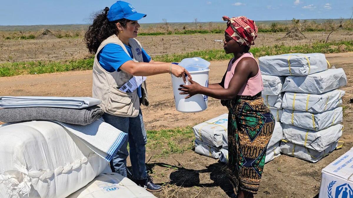 A woman wearing a blue UNHCR shirt and cap hands out provisions to a woman dressed in a pink blouse, colorful pants, and a red headscarf. Behind them, there is a dirt field with patches of green grass.