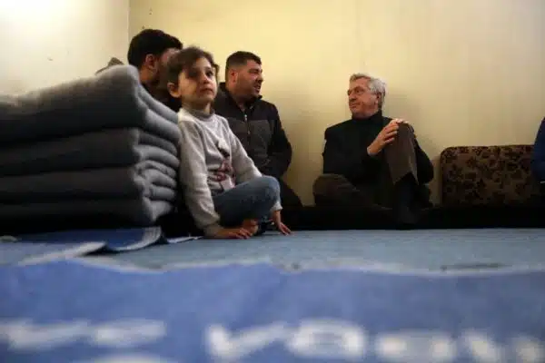 A little girl sits cross-legged on the floor next to a stack of folded blankets as three older pictured in the background men converse with each other.