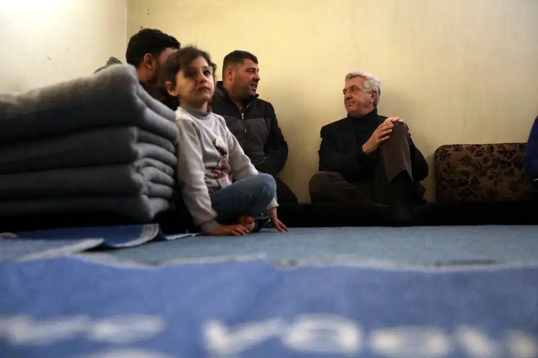 Une petite fille est assise les jambes croisées sur le sol, à côté d'une pile de couvertures pliées, tandis que trois hommes plus âgés, représentés à l'arrière-plan, discutent entre eux. 