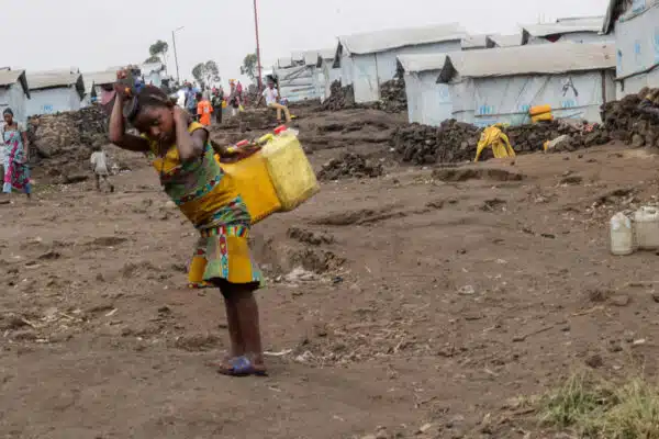 A young girl carries two large yellow plastic water jars on her back that are strapped to her forehead . In the background a row of white shelters with UNHCR logos on plastic sheeting is visible.