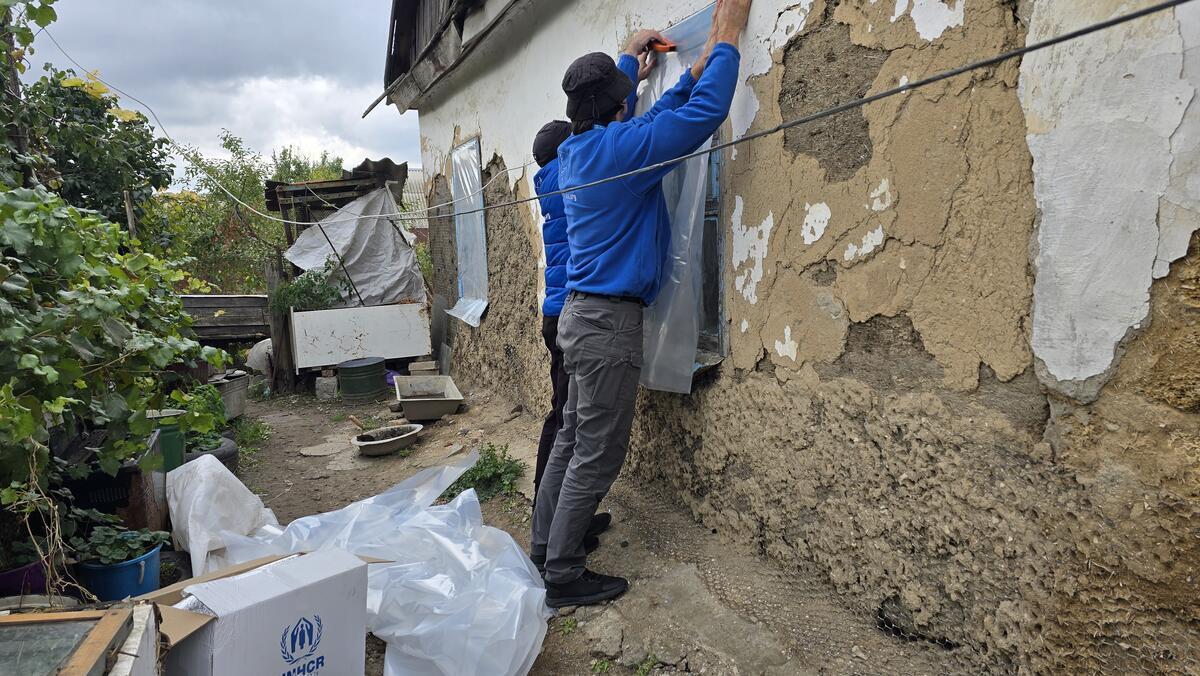 A man is hanging a plastic sheet over a window to protect it from the wind and bad weather. There is a damaged home around him.