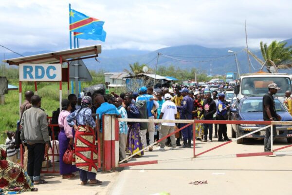 A crowd of people stands by a closed gate barrier on a road as two blue flags with a diagonal red line flutter in the wind. Forest covered mountains with clouds over them can be seen in the distance.