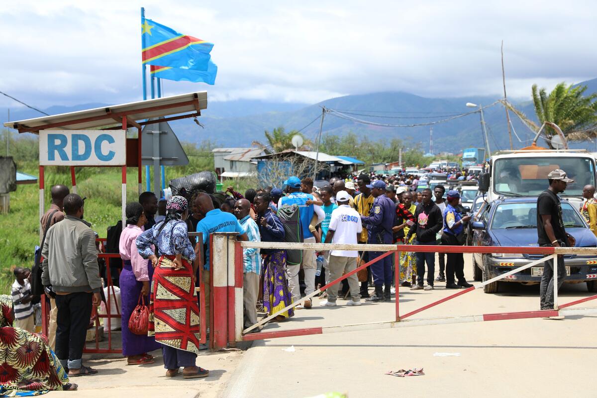 A crowd of people stands by a closed gate barrier on a road as two blue flags with a diagonal red line flutter in the wind. Forest covered mountains with clouds over them can be seen in the distance. 