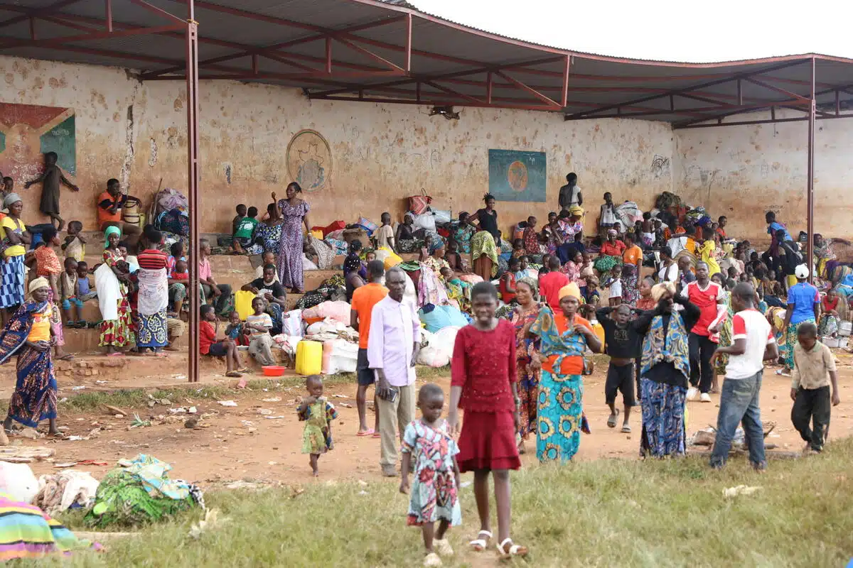 A large group of people including women and children are gathered under an open-air structure with an awning to protect from the elements.